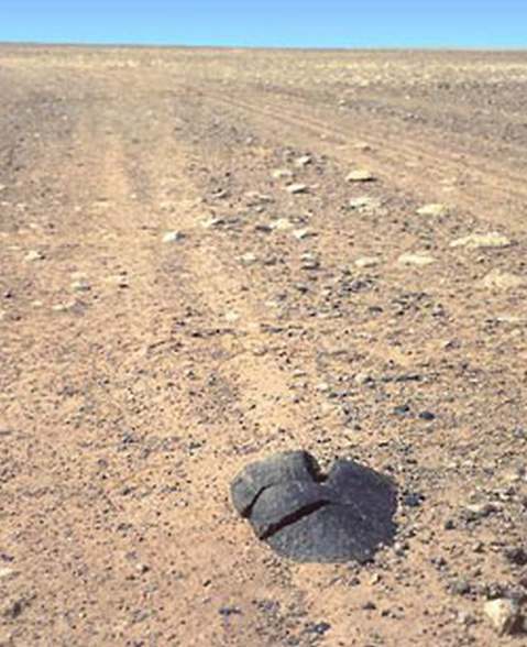 Черный каменный метеорит массой 30 кг, хорошо заметный на светлом фоне каменистого плато в ливийской Сахаре, случайно попал под колесо автомобиля французской экспедиции и развалился на три части, что сильно снизило его рыночную стоимость. Фото: © SaharaMet / R. & R.Pelisson  