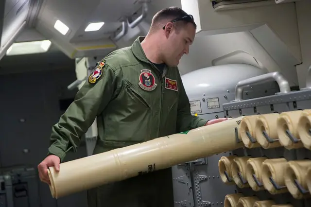Офіцер ВМС США завантажує гідроакустичний буй на стелаж для використання під час пошуків рейсу MH370. Він одягнений у армійський зелений комбінезон та має зачіску короткої стрижки. Гідроакустичний буй, який він завантажує, має циліндричну форму та бежевого кольору. Здається, він знаходиться в літаку.