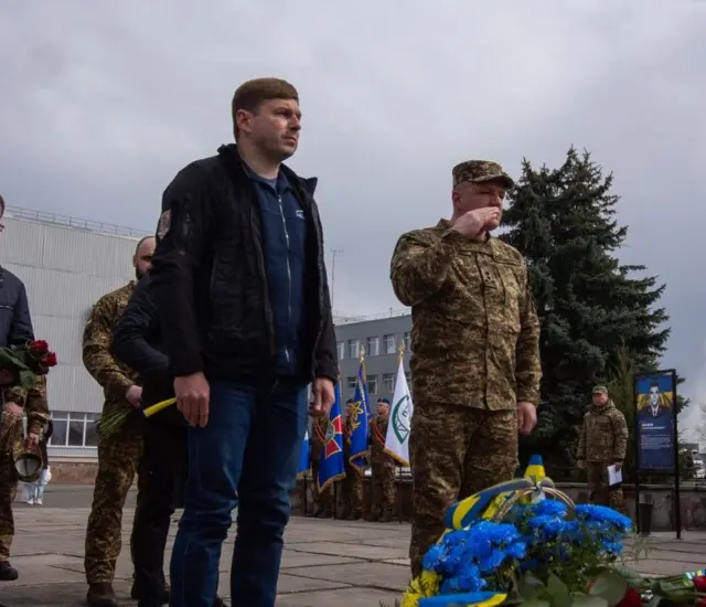 Сергій Тараканов у темно синьому светрі стоїть перед меморіалом з квітами 