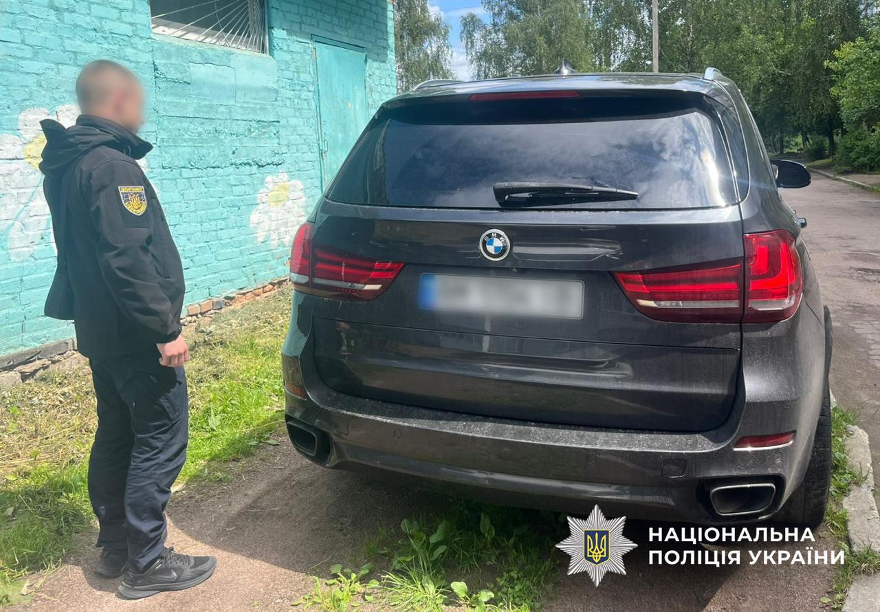 На зображенні показано задню частину сучасного кросовера BMW темного кольору, що стоїть на узбіччі дороги біля будівлі. Ліворуч від автомобіля спиною до глядача стоїть чоловік у темному одязі — куртці з нашивкою-шевроном на правому плечі та штанах. Його обличчя повернуте у профіль та розмито. Чоловік дивиться у бік стіни будівлі. Сама будівля зліва побудована з цегли та пофарбована у світло-бірюзовий колір, на ній видно намальовані білі ромашки. Автомобіль припаркований частково на ґрунті з травою. На авто помітно фірмовий значок BMW, а автомобільний номерний знак розмито з міркувань конфіденційності. На задньому плані вздовж дороги ростуть зелені дерева під яскравим денним небом. У нижньому правому кутку фотографії розміщено білий логотип Національної поліції України з восьмикутною зіркою, малим гербом України та написом Національна поліція України.