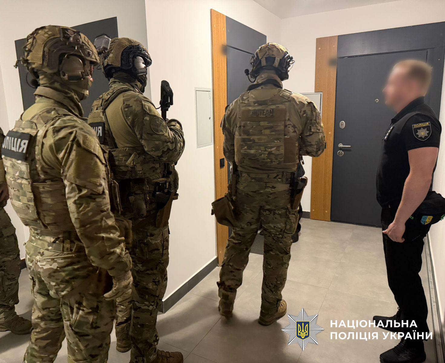 Світлина з коридору будівлі, де стоять четверо чоловіків. Праворуч профільним боком зображено чоловіка у чорній футболці-поло з шевроном Поліція на рукаві та у чорних штанах, його обличчя розмите. Навпроти нього спиною та напівпрофілем до глядача стоять троє чоловіків у камуфляжній військовій формі, захисних шоломах, тактичних навушниках та бронежилетах із кобурами на поясах. На спині та плечі одного з бійців у камуфляжі видно нашивку з написом Поліція. На задньому плані розташовані сірі двері з дерев'яною лиштвою та білі стіни. Підлога викладена світлою квадратною плиткою. У правому нижньому кутку поверх фотографії накладено офіційний логотип Національної поліції України у вигляді білої восьмикутної зірки з тризубом на синьому тлі та білий напис Національна поліція України.