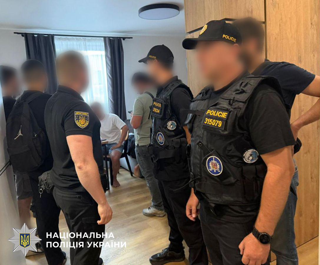Світлина з інтер'єру кімнати, де перебуває група чоловіків, обличчя яких розмиті. На передньому плані праворуч боком до об'єктива стоїть чоловік у темній формі, кепці з написом POLICIE та тактичному жилеті з нашивкою POLICIE і круглими шевронами. Поруч із ним та позаду стоять ще двоє чоловіків у аналогічній формі та кепках із написом POLICIE. Ліворуч спиною до глядача стоїть чоловік у чорній футболці-поло з жовтим круглим шевроном Кіберполіції на рукаві. На задньому плані в глибині кімнати видно чоловіка у світлій футболці та джинсах, а також чоловіка у білій футболці та шортах, який сидить на стільці біля вікна з темними шторами. У кімнаті видно дерев'яну підлогу та меблі. У лівому нижньому кутку поверх фотографії накладено офіційний білий логотип-зірку з гербом-тризубом на синьому тлі та білий напис Національна поліція України.