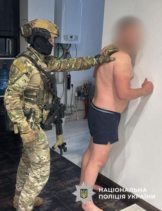 На зображенні зафіксовано момент затримання чоловіка правоохоронцем у приміщенні. Ліворуч стоїть спецпризначенець у повній бойовій екіпіровці: камуфляжній формі, тактичному шоломі, навушниках, балаклаві, бронежилеті та рукавицях. Через плече у нього тримається автоматична зброя. Спецпризначенець поклав руку на плече затриманому, контролюючи його рухи. Праворуч, обличчям до білої стіни, стоїть затриманий чоловік міцної статури. Він босий, одягнений лише у темні шорти, його обличчя розмито з міркувань конфіденційності. Чоловік спирається руками на стіну. Події відбуваються в кімнаті з темною підлогою, на задньому плані видно частину кухонних меблів та побутову техніку, зокрема газовий котел чи бойлер на стіні. У нижній частині кадру по центру розміщено офіційний логотип Національної поліції України із зображенням восьмикутної зірки, тризуба та відповідного напису.