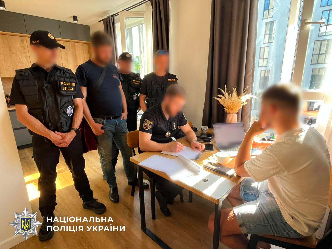 Світлина з інтер'єру світлої кімнати, де за дерев'яним столом біля вікна сидять двоє чоловіків, а поруч стоять ще кілька людей. Обличчя всіх присутніх розмиті. Праворуч спиною до глядача сидить чоловік у білій футболці та джинсових шортах, перед яким на столі стоїть відкритий ноутбук і біла чашка. Навпроти нього за столом сидить чоловік у темній формі з шевроном Нацполіції та робить записи у документах. Ліворуч від них стоять четверо чоловіків: один у цивільному одязі — синій футболці та джинсах, а троє інших — у темній тактичній формі з написом POLICIE та номерами на грудях. На столі також лежать папери, мобільний телефон і стоїть ваза з сухоцвітом. Через велике вікно на підлогу падає сонячне світло. У лівому нижньому кутку зображення розміщено білий логотип-зірку з тризубом та напис Національна поліція України.
