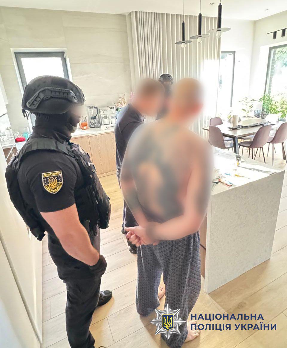 Світлина з інтер'єру світлої кухні-вітальні, де перебуває кілька чоловіків, обличчя яких розмиті. На передньому плані ліворуч боком до об'єктива стоїть чоловік у чорному тактичному одязі, шоломі та бронежилеті, на рукаві якого є жовтий круглий шеврон Департаменту кіберполіції. Поруч із ним спиною до глядача стоїть босоногий чоловік без сорочки у сірих картованих штанах, який тримає руки схрещеними за спиною. На його спині та плечах помітні татуювання. На задньому плані стоїть ще один чоловік у темному одязі, видно кухонну стійку з кавомашиною, а також обідній стіл із чотирма стільцями під підвісними світильниками. У правому нижньому кутку поверх фотографії накладено офіційний білий логотип-зірку з гербом-тризубом на синьому тлі та синій напис Національна поліція України.