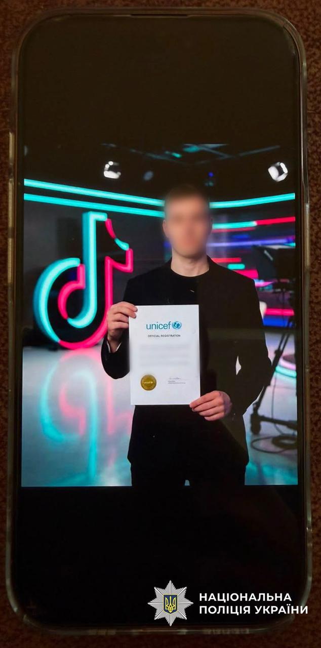 На зображенні показано вертикальний екран смартфона, на якому відкрита фотографія чоловіка. Чоловік одягнений у темний піджак та чорну футболку, його обличчя розмите. Він стоїть у приміщенні та тримає обома руками перед собою білий аркуш паперу формату А4. У верхній частині документа видно синій логотип UNICEF та напис OFFICIAL REGISTRATION, а в лівому нижньому куті аркуша — круглу золотисту печатку або наклейку. Позаду чоловіка розташована велика підсвічена неонова емблема соціальної мережі TikTok у блакитних та рожевих кольорах. У нижній частині екрана смартфона поверх фотографії накладено логотип Національної поліції України — срібну восьмикутну зірку з тризубом та білий напис НАЦІОНАЛЬНА ПОЛІЦІЯ УКРАЇНИ.