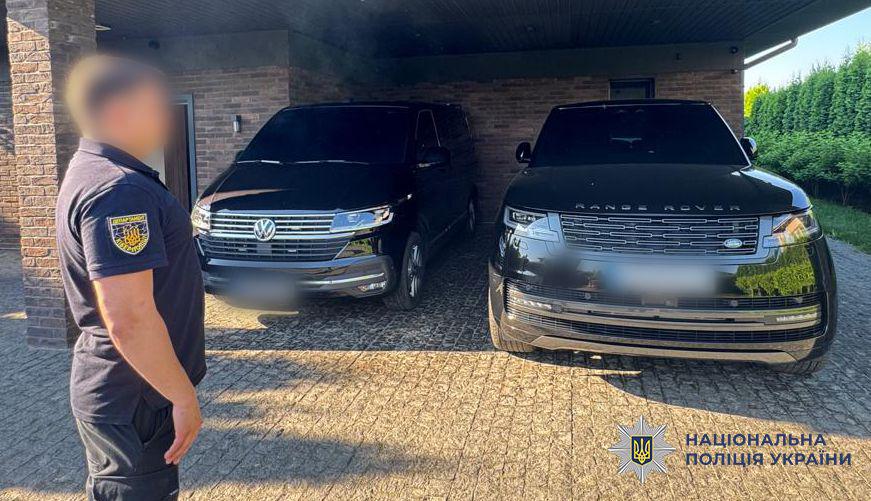 Фотографія на вулиці біля приватного будинку з бруківкою, де під навісом припарковані два автомобілі чорного кольору передом до об'єктива. Ліворуч стоїть мінівен марки Volkswagen з хромованою решіткою радіатора та фірмовим круглим логотипом, а праворуч — великий позашляховик марки Range Rover із відповідним написом на капоті. Номерні знаки обох транспортних засобів розмиті. На передньому плані ліворуч у профіль стоїть чоловік у темній футболці-поло з жовтим круглим шевроном Кіберполіції на рукаві та темних штанах, його обличчя розмите. Стіни будинку викладені коричневою цеглою, а праворуч на задньому плані видно зелені туї та газон. У правому нижньому кутку поверх фотографії накладено офіційний білий логотип-зірку з гербом-тризубом на синьому тлі та синій напис Національна поліція України.