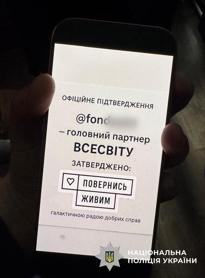 На зображенні людина тримає в руці смартфон із яскравим білим екраном у темному приміщенні. На екрані телефона відображається текст чорними літерами на білому тлі. Зверху написано: офіційне підтвердження, нижче йде нікнейм інстаграм-акаунту, який частково розмито, а далі текст: — головний партнер ВСЕСВІТУ. ЗАТВЕРДЖЕНО: лаконічний логотип фонду Повернись живим у вигляді трьох прямокутних блоків із серцем та словами повернусь живим. Знизу дрібним шрифтом додано: галактичною радою добрих справ. У правому нижньому кутку самого зображення розміщено офіційний логотип Національної поліції України — восьмикутна зірка з малим гербом по центру та напис Національна поліція України.