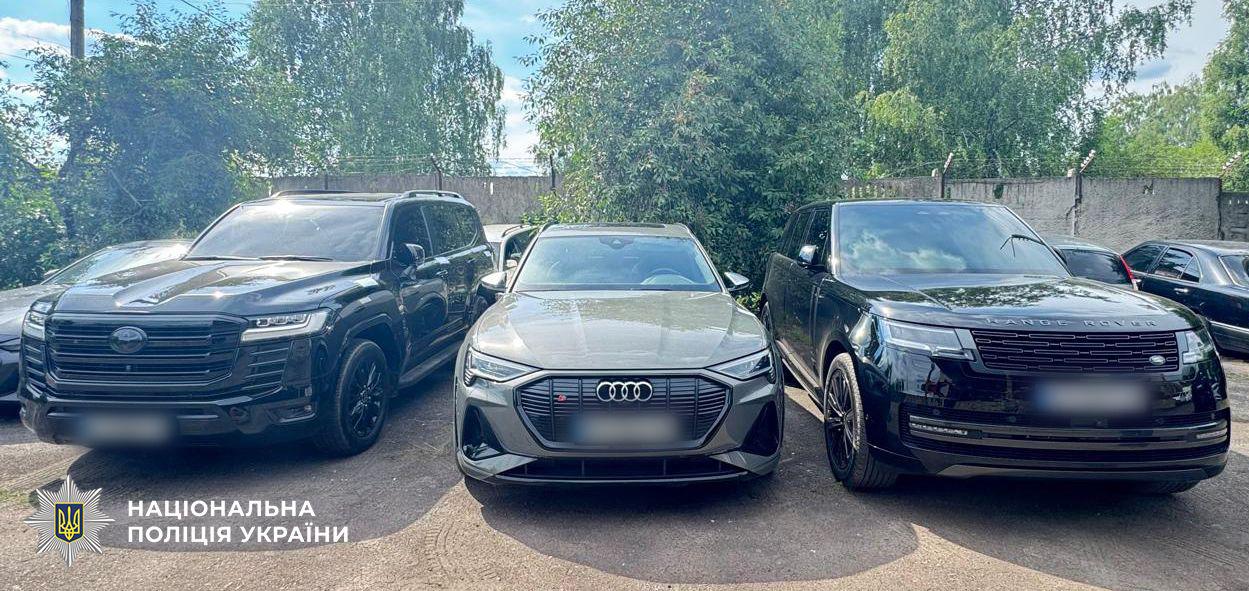 Фотографія трьох сучасних легкових автомобілів, які припарковані в один ряд на відкритому майданчику передом до об'єктива. Зліва стоїть великий позашляховик чорного кольору, по центру — сірий кросовер марки Audi з характерним фірмовим логотипом із чотирьох кілець на решітці радіатора, а справа — чорний позашляховик марки Range Rover з відповідним написом на капоті. Номерні знаки всіх трьох автівок розмиті. На задньому плані за автомобілями видно високий бетонний паркан із колючим дротом зверху, а за ним ростуть густі зелені дерева під світлим небом із легкими хмарами. У лівому нижньому кутку поверх фотографії накладено офіційний білий логотип-зірку з гербом-тризубом на синьому тлі та білий напис Національна поліція України.